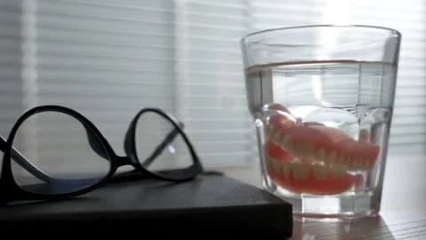 False teeth in glass of water on table with notebook and glasses, close-up Stock Footage 288856121