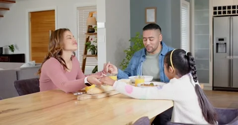 Family linking hands at dining table with cereal, mother raising arms toasting 스톡 동영상 338167596