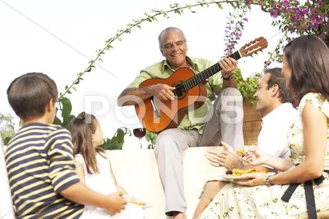 Family Sitting Outdoors Listening To Man Playing Guitar