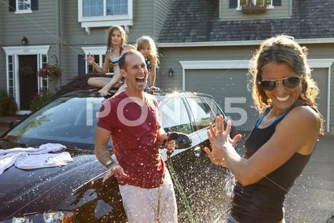 A Family Washes Their Car In The Driveway Of Their Home On A Sunny Summer