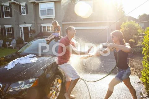 A Family Washes Their Car In The Driveway Of Their Home On A Sunny Summer