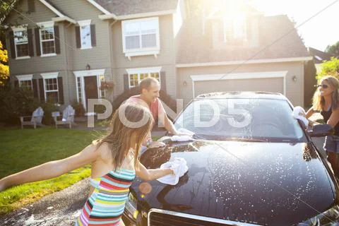 Family Washing Their Car In The Driveway Of Their Home On A Sunny Summer