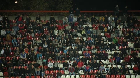Fans crowd sitting on stadium seats, watching football or soccer game at night Stock Footage
