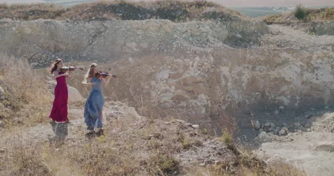 Far view of two elegant musicians in dresses play violin among cliff in summer Stock Footage 136074235