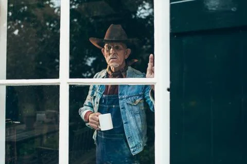 Farmer with beard and hat holding cup of coffee looking out window of old far Foto stock