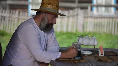 Farmer, man with a beard in a straw hat, sits at a table and drinks dark beer Stock Footage 81633761