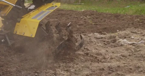 Farmer using a yellow motorized tiller to plow the land in a garden, loosening 스톡 동영상 333759932