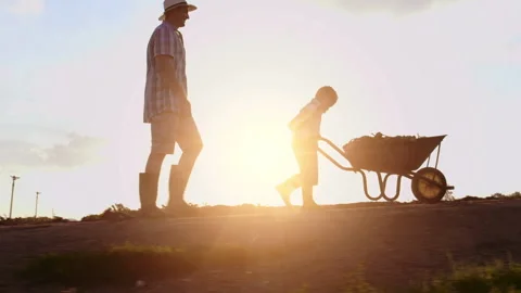 Farming father helping son push wheelbarrow up hill on their farm at sunset. Stock Footage 199631529