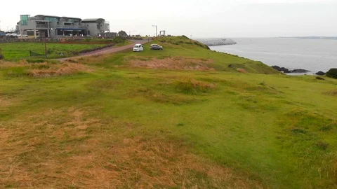 Fast low drone shot over grass near the sea going up over a clifftop restaurant Video stock 91092436
