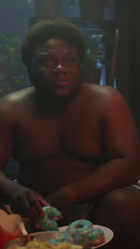 Fat Black Man Eating Donut and Laughing at TV Show in Evening at Home Stock Footage 276880572