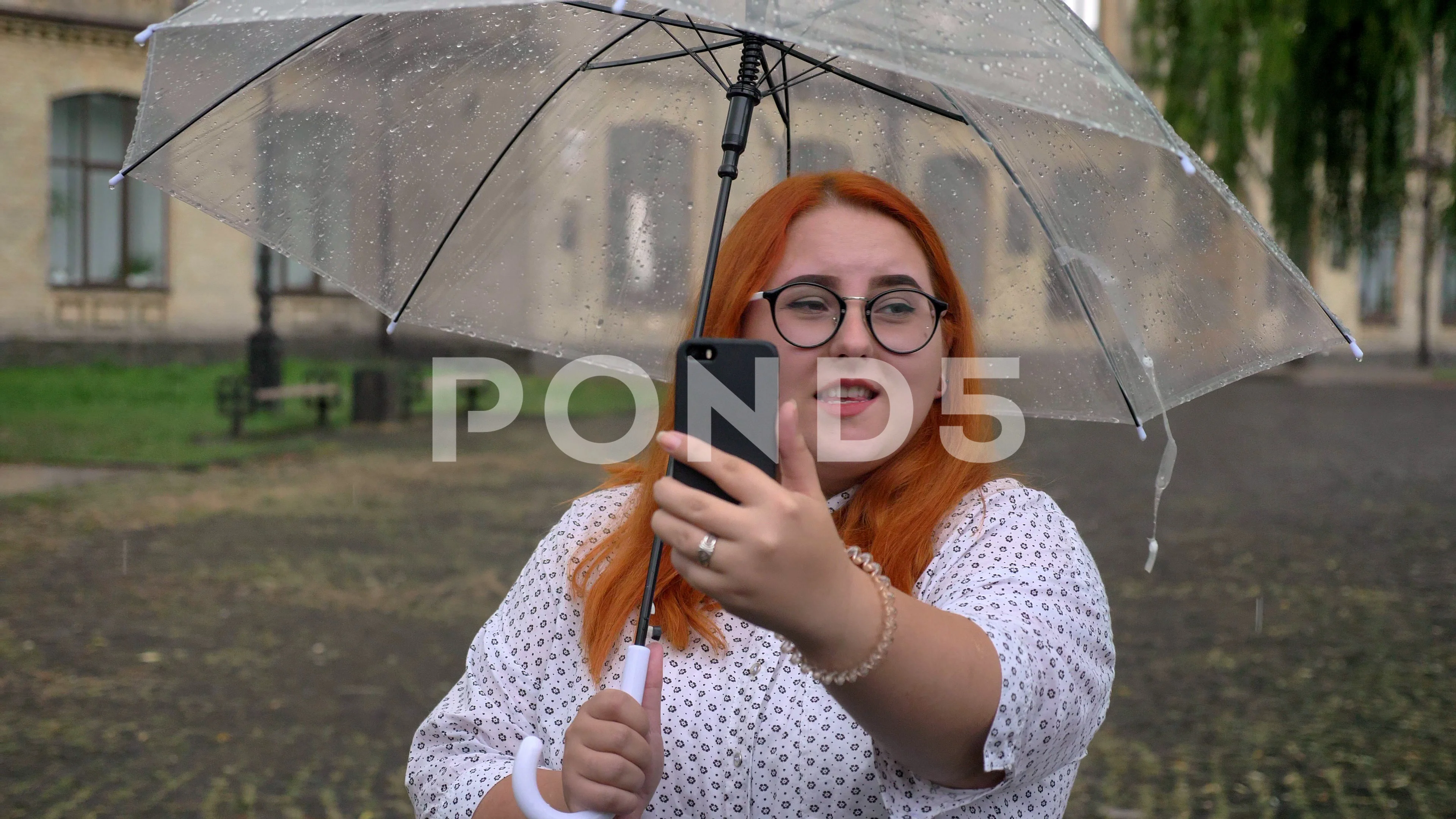 Fat ginger girl with glasses is talking in videochat on smartphone in park  in