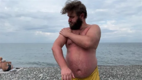 Fat man with beard, big belly in yellow shorts applies cream to his body Stock Footage 310397053