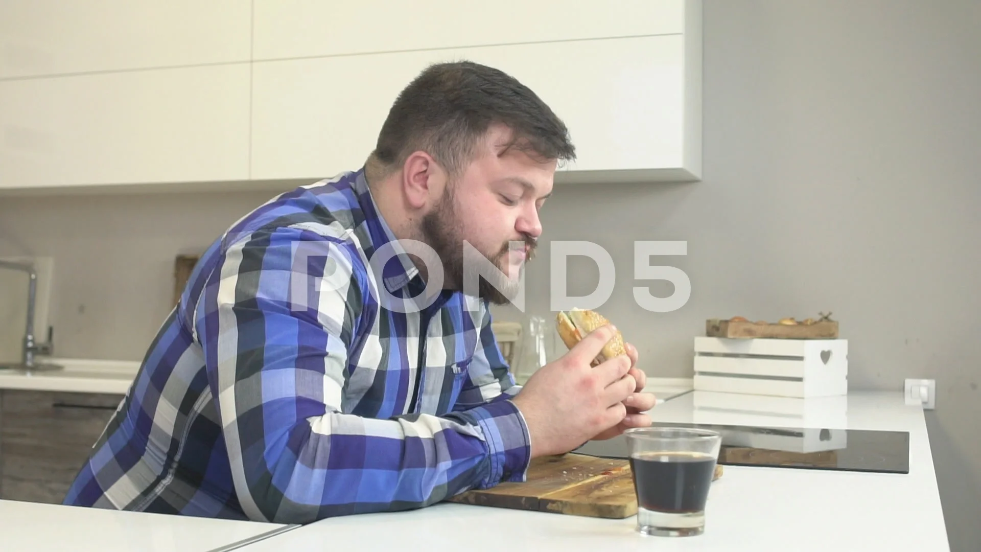 Fat man with a beard in a plaid shirt eats fast food cheeseburger and washes