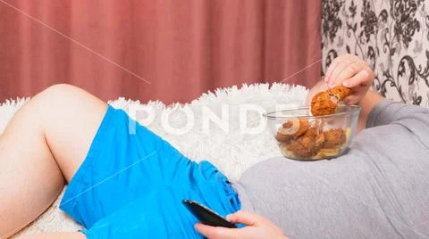 Fat man with a big belly is lying on the couch with fast food and is bored in Stock Photos