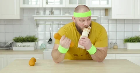 Fat man eating danar sitting at table and doing exercise for hand with dumbbell. Stock Footage 149070334