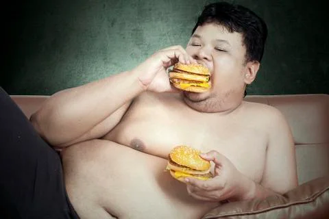 Fat man eating two hamburgers while watching tv at home Stock Photos