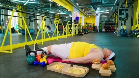 The fat man is exercising in the gym and looking at the burger and pizza. Obesit Stock Footage 244240231