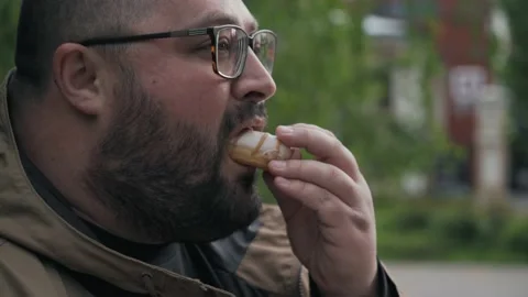 Fat man in glasses eats donut and drinks coffee on street, close up portrait Stock Footage 130748585