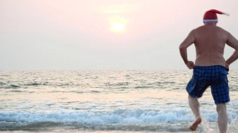Fat man in a red Santa hat runs funny holding shorts and dives into sunset sea. Stock Footage 117726392