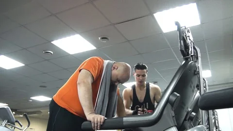 Fat man is very tired on the treadmill. His young coach motivates him Stock Footage