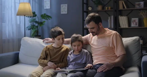 Father and children sitting on the sofa in the evening. Little girl reading book Stock Footage 274601787