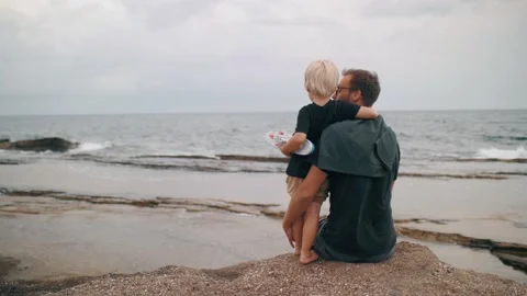 Father and son hugging talking together contemplating dramatic sea wave beach Stock Footage 225875957