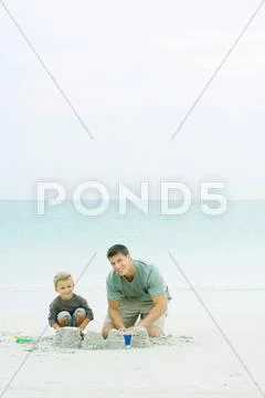 Father And Son Making Sand Castles At The Beach, Smiling At Camera
