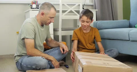 Father and son open cardboard box on floor together. Focused father teaches boy Stock Footage 328706805