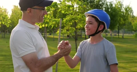 Father and son share moment in sunny park. Boy wears helmet and holds scooter Stock Footage 346753398