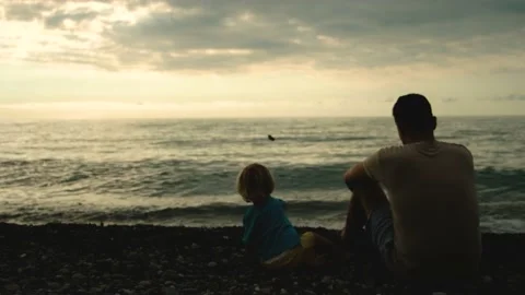 Father and son sit back on the beach and throw stones into the sea at the sunset Video stock 200807401