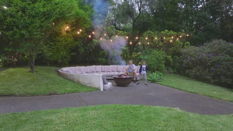 Father and son spending time by fire pit in backyard with string lights at dusk. 스톡 동영상 338369190