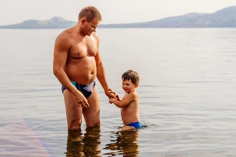 Father and son in swimming trunks stand in the lake, holding hands Stock Photos