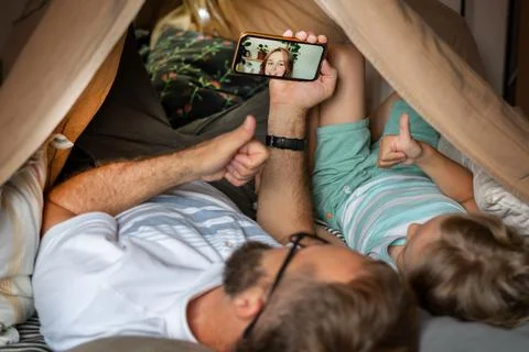 Father and son using mobile phone while lying in children's tent, making video Stock Photos