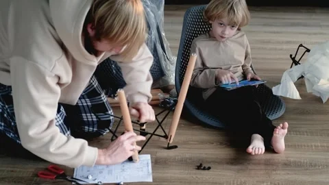 Father assembles chair while son sits nearby playing on tablet Stock Footage 305947775