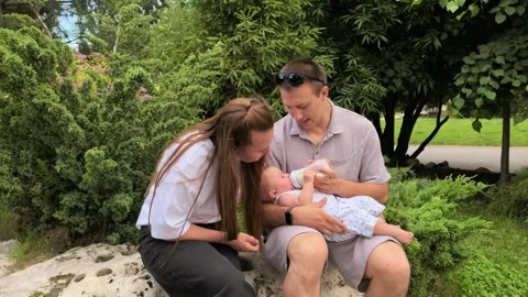 Father bottle feeds an infant while the mother watches outdoors in a park Video stock 340189825
