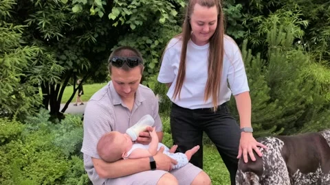 Father bottle feeds an infant while sitting with a woman and dog in a park Stock Footage 340189838