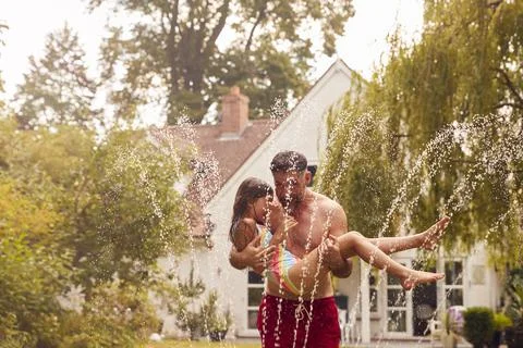 Father Carries Daughter Through Water From Garden Sprinkler Having Fun Wearing Stock Photos