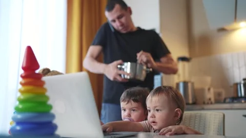 Father cooking in the kitchen remote work. baby twins playing with laptop in the Stock Footage 268574402