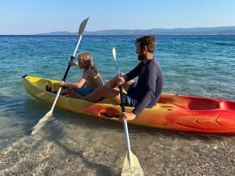 Father guiding young boy while paddling kayak in calm coastal water Stock Photos