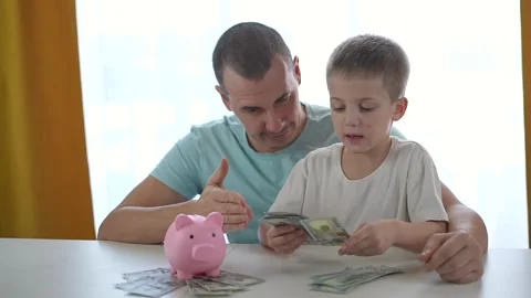 Father helps boy count dollar bills during home finance session. Piggybank Stock Footage 320113888