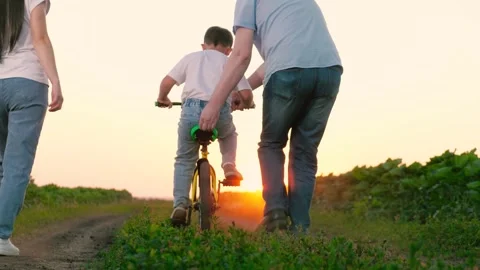 Father holds bicycle seat supporting child pedaling near walking mother at Stock Footage 260189689