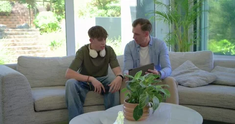Father opening tablet, showing content and guiding teenage son scrolling on sofa Stock Footage 336784521