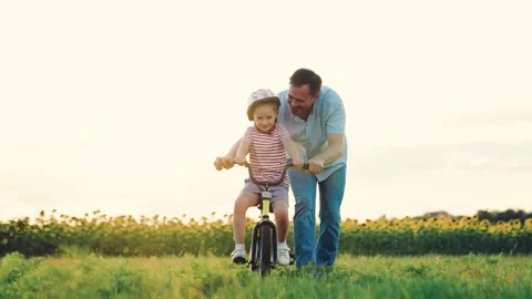 Father pulls bicycle while little girl rides vehicle in summer rural meadow Stock Footage 255318944