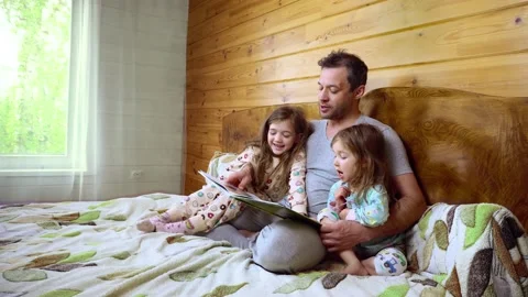 Father sits on the bed with his cute little daughtersin and reads the book. Stock Footage 164902893