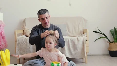 A father sits on the floor arranging his daughter's hair as she plays with Vídeos de archivo 336460046