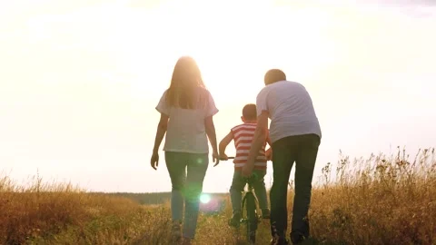 Father supports son learning to ride bicycle resting with family at countryside Stock Footage 246842904