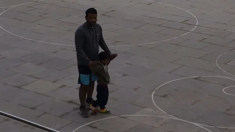 A father teaches his son roller blades at a public playground in India Stock Footage 83459309