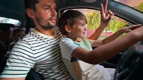 Father teaching his young son how to drive car on his lap. Showing peace sign Stock Footage 250359265