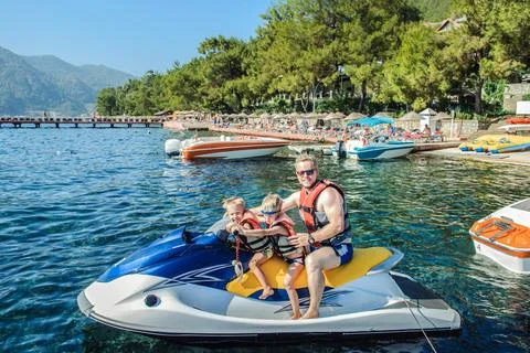 Father with two small sons wearing life jackets is seated on a jet ski near.. Stock Photos
