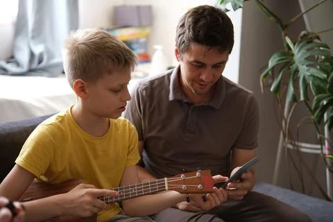 Father using smartphone teaching son to play ukulele at home Stock Photos